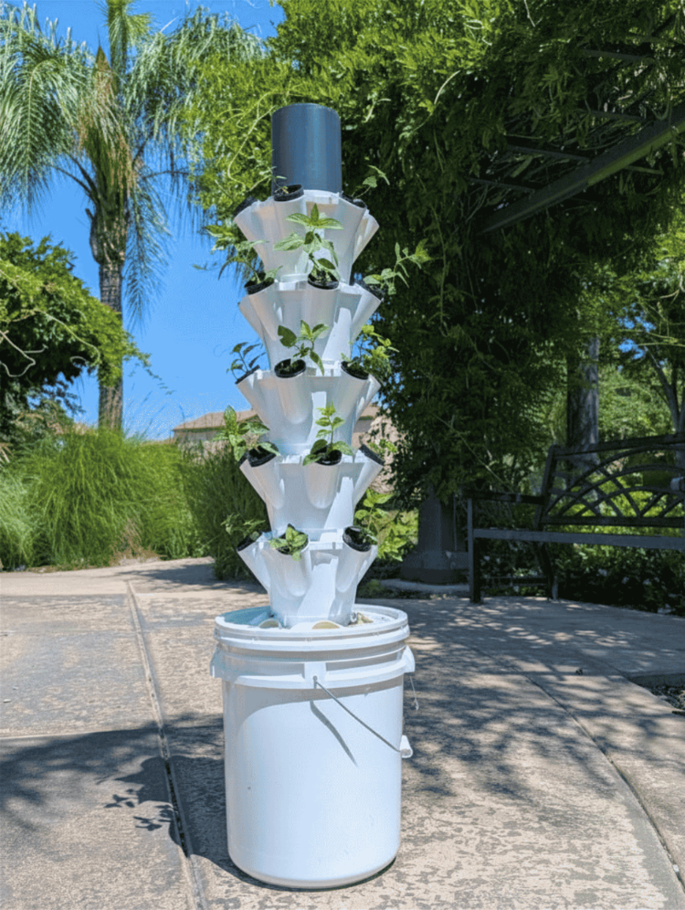 A vertical tiered hydroponic garden tower made of white plastic cups and pots, growing small green plants, set outdoors on a concrete patio with trees and a bench in the background.